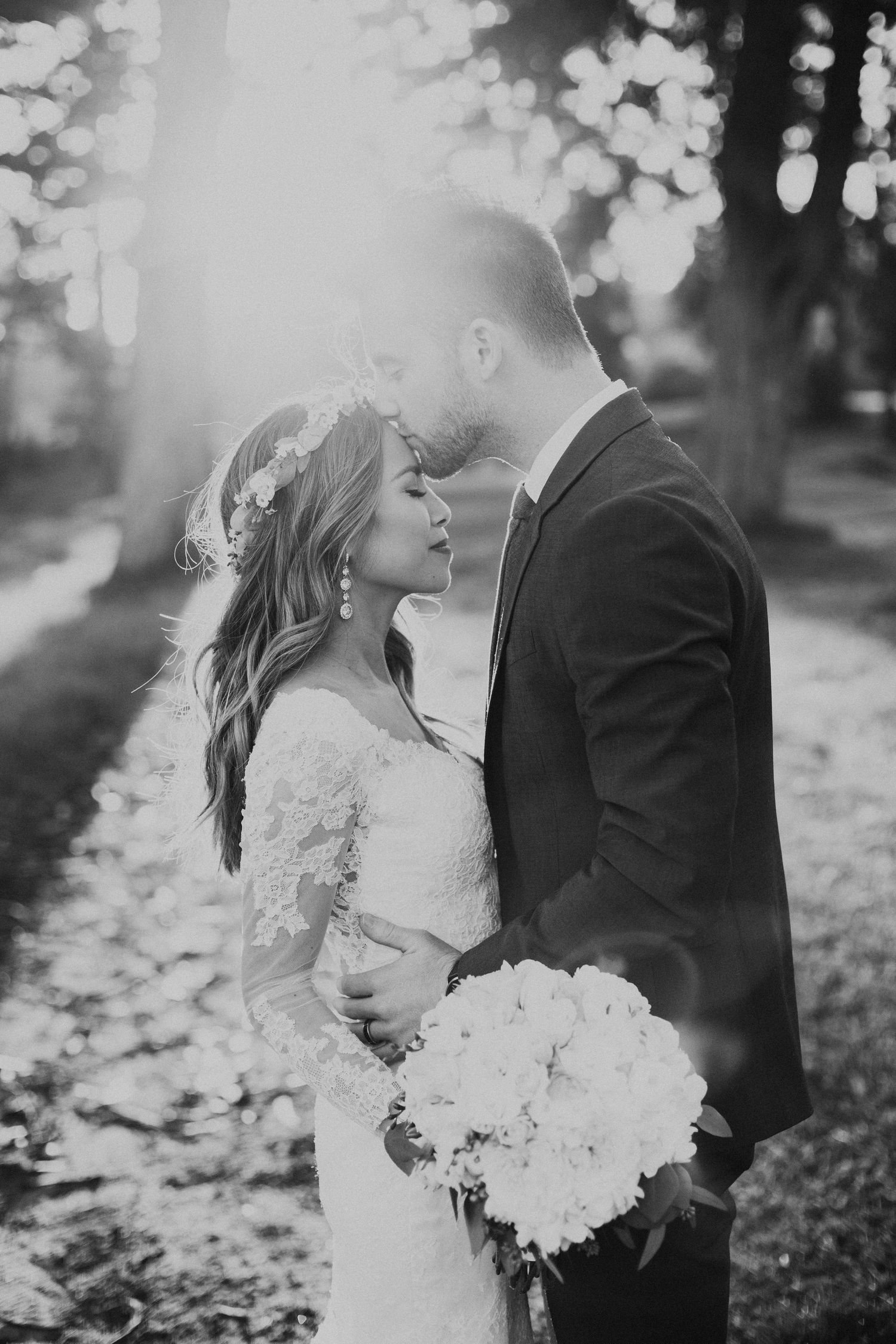 Romantic black and white wedding portrait with couple embracing in sunlit forest setting.