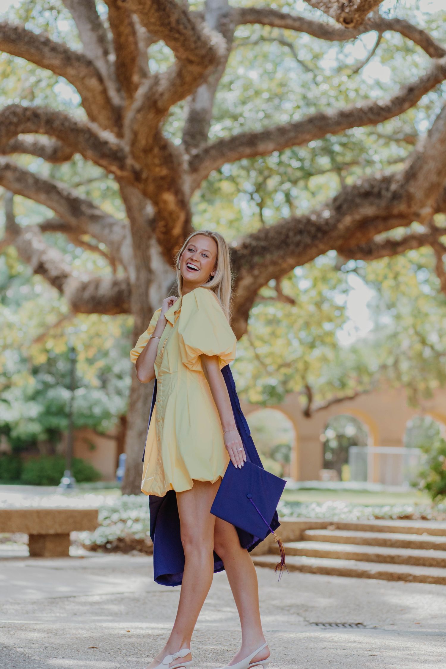 A joyful graduate in a yellow dress twirls under a large oak tree on campus.