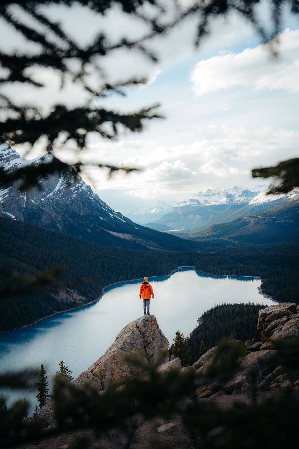 Hiker in orange jacket stands on rocky overlook above turquoise alpine lake surrounded by snow-capped mountains.