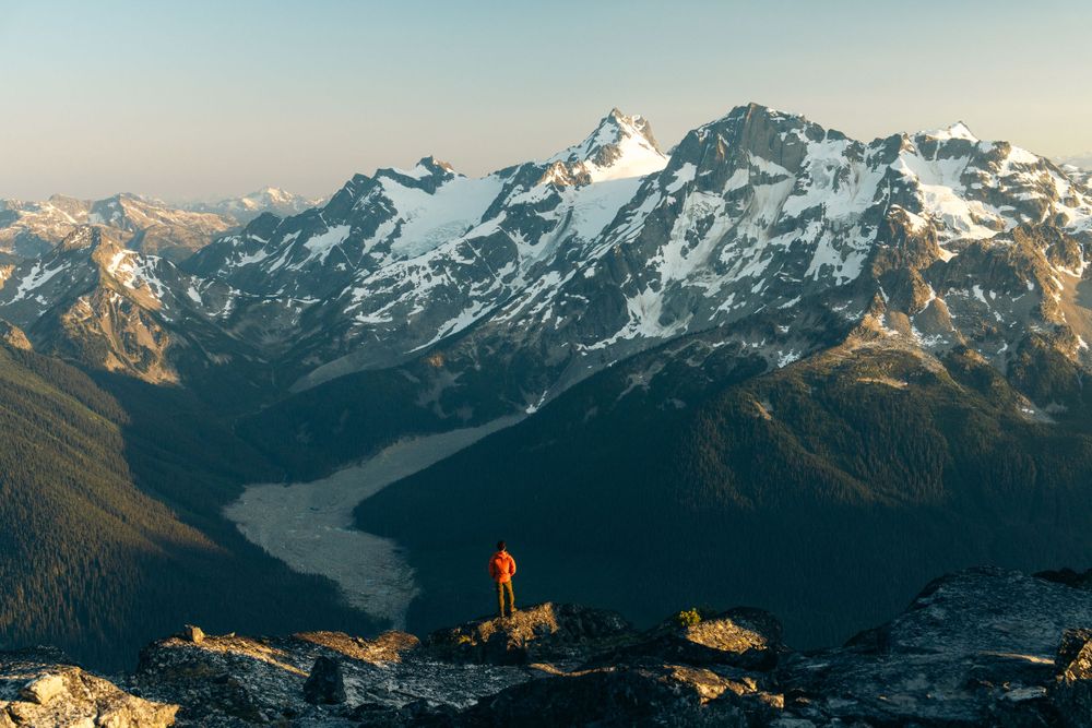 Distant figure in orange stands on mountain ridge overlooking snow-capped alpine valley.