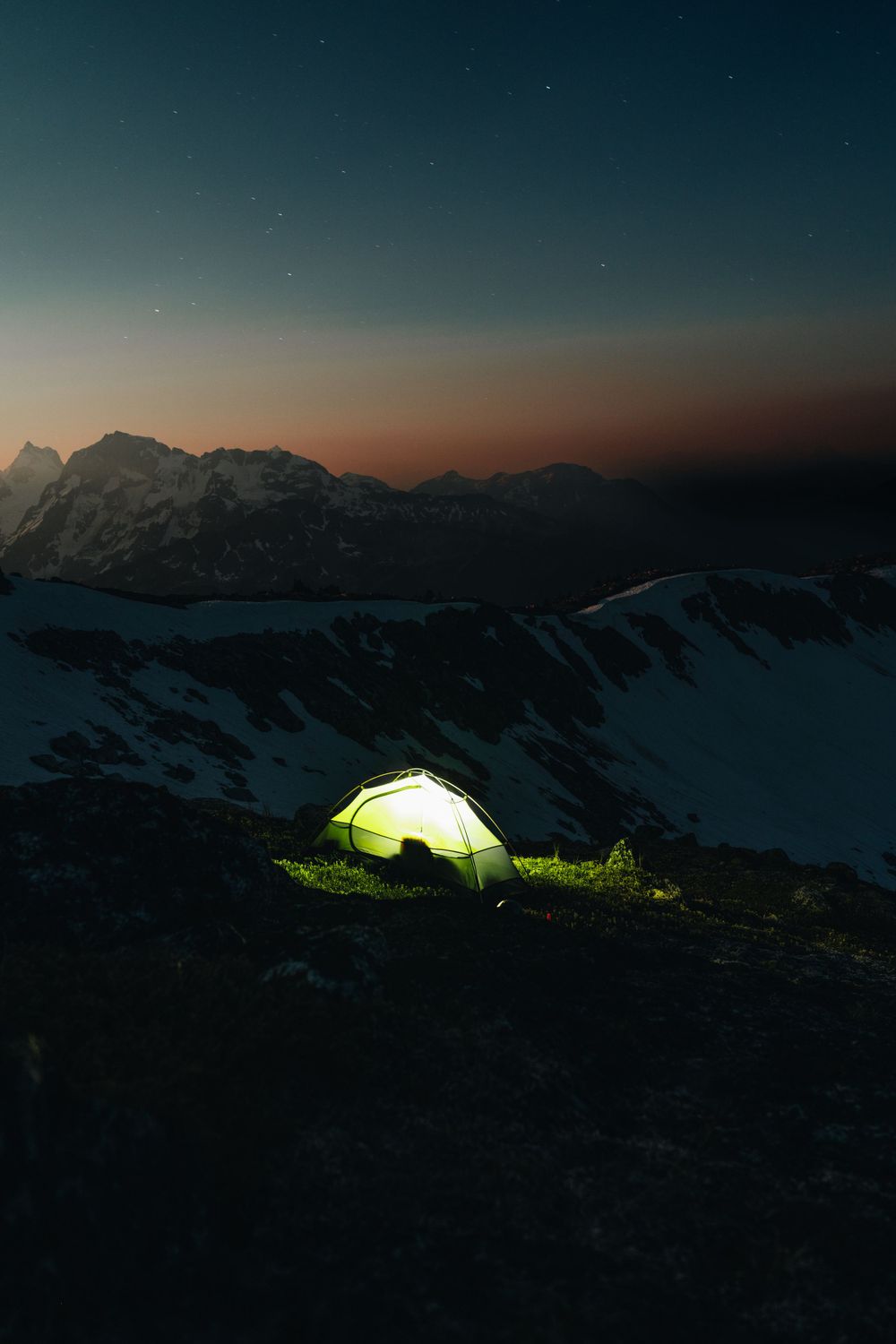 Glowing tent illuminated against starry night sky with snowy mountain backdrop.