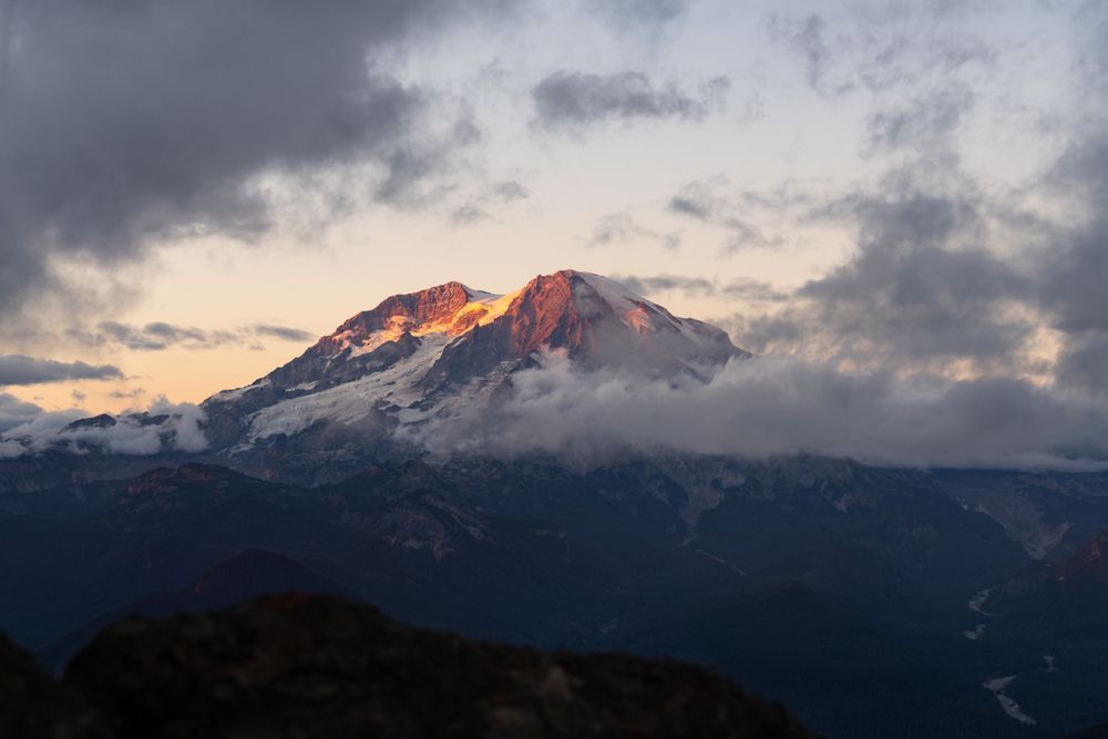 Snow-dusted mountain peak glows pink during sunset with dramatic cloudy sky.
