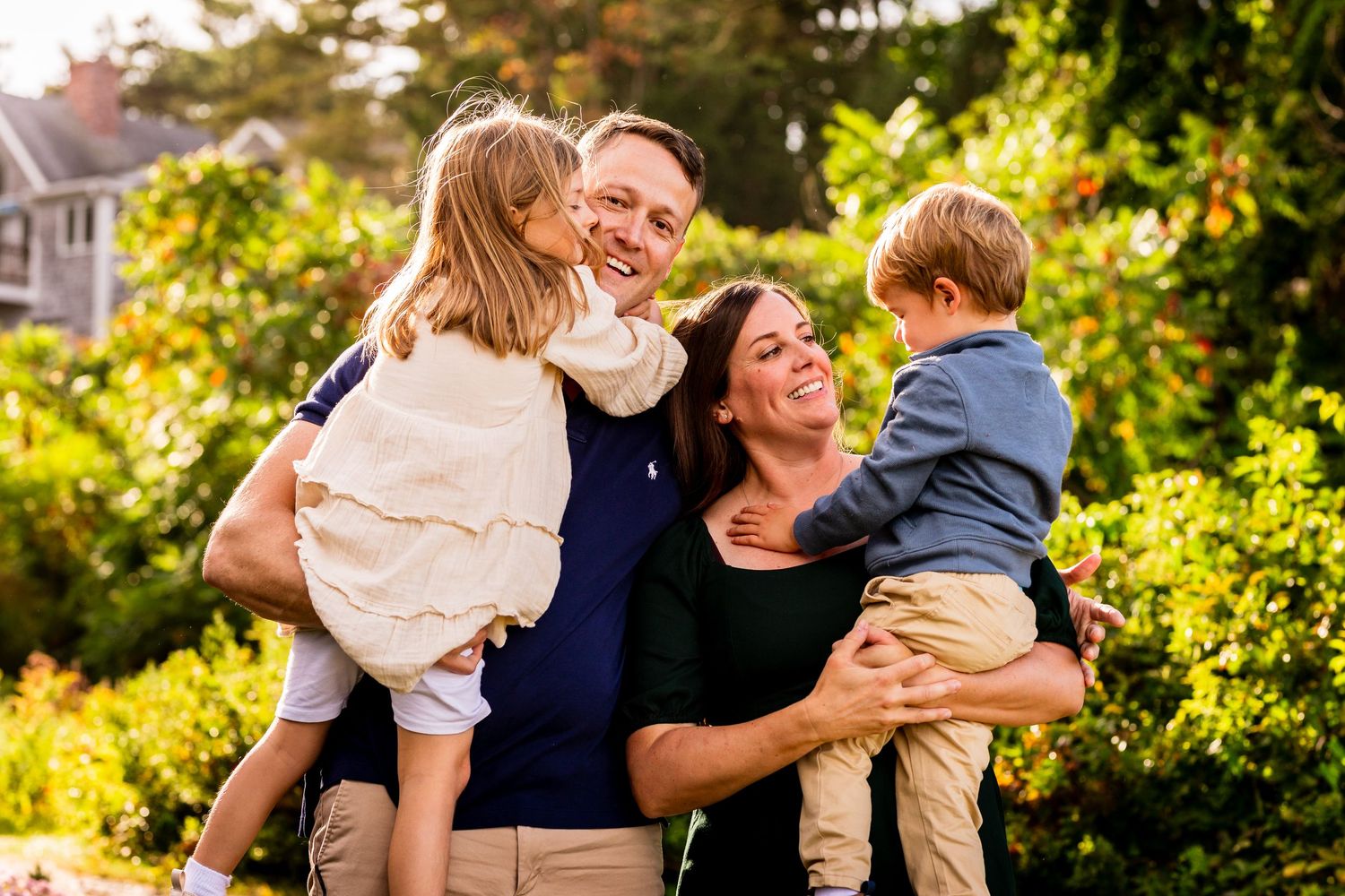 A loving family embraces and laughs together outdoors in a sunlit garden setting during golden hour.