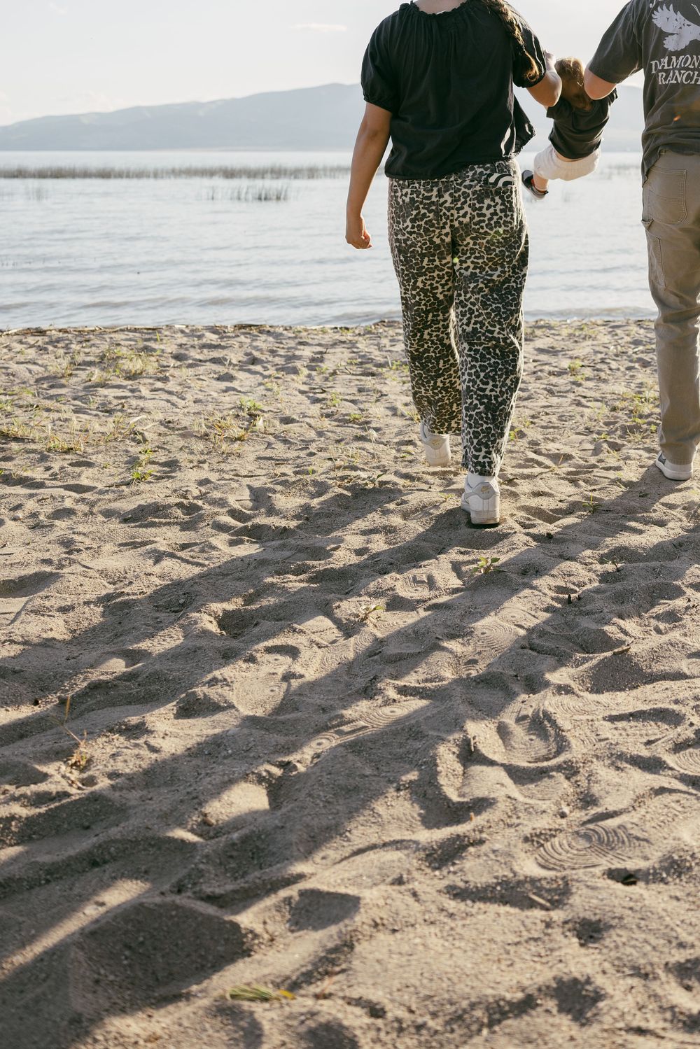Two people walking on a sandy beach with long shadows, wearing casual clothes including leopard print pants.