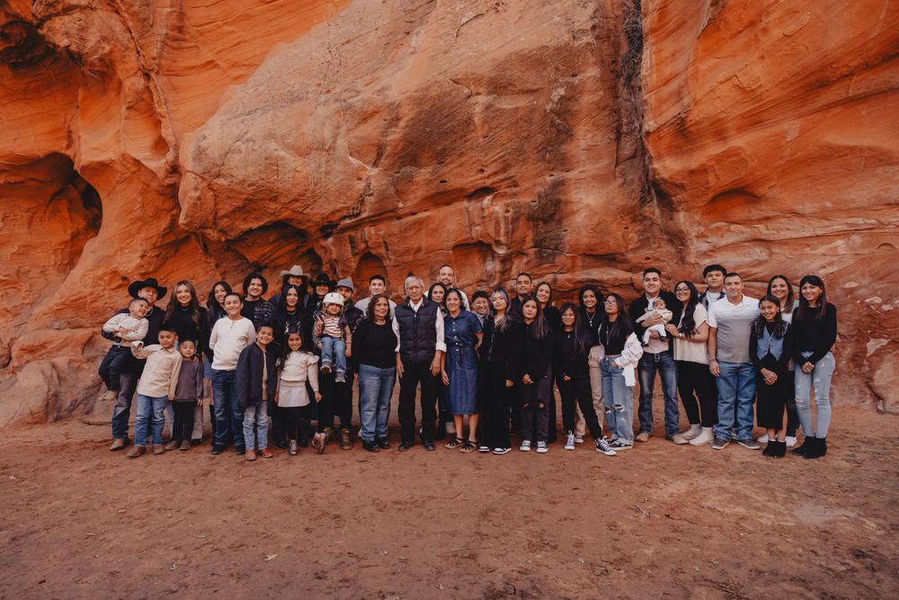 Large group photo of people gathered in front of towering red rock cliffs in outdoor setting.