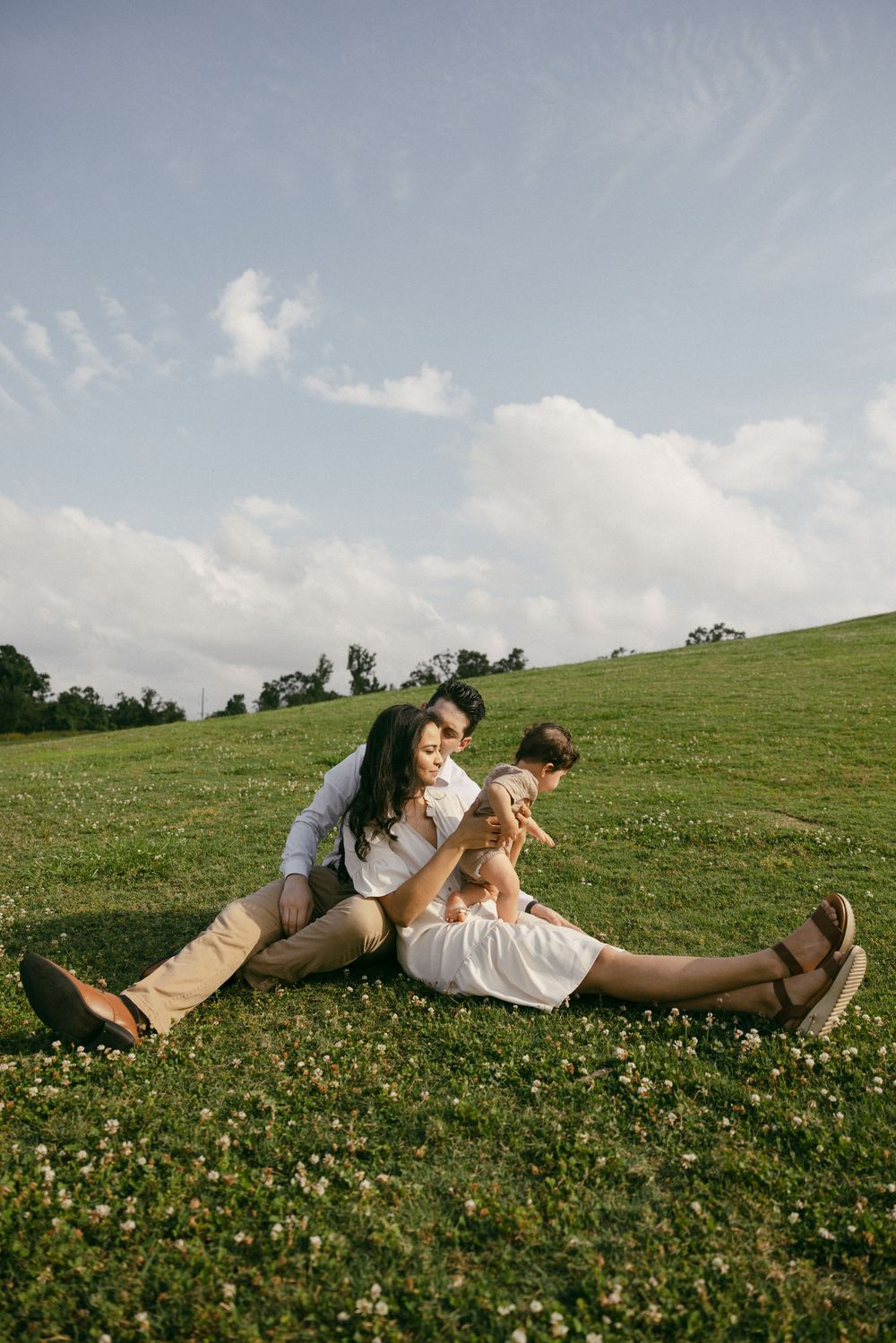Two figures relax on a sunny hillside covered in wildflowers with blue sky and white clouds overhead.