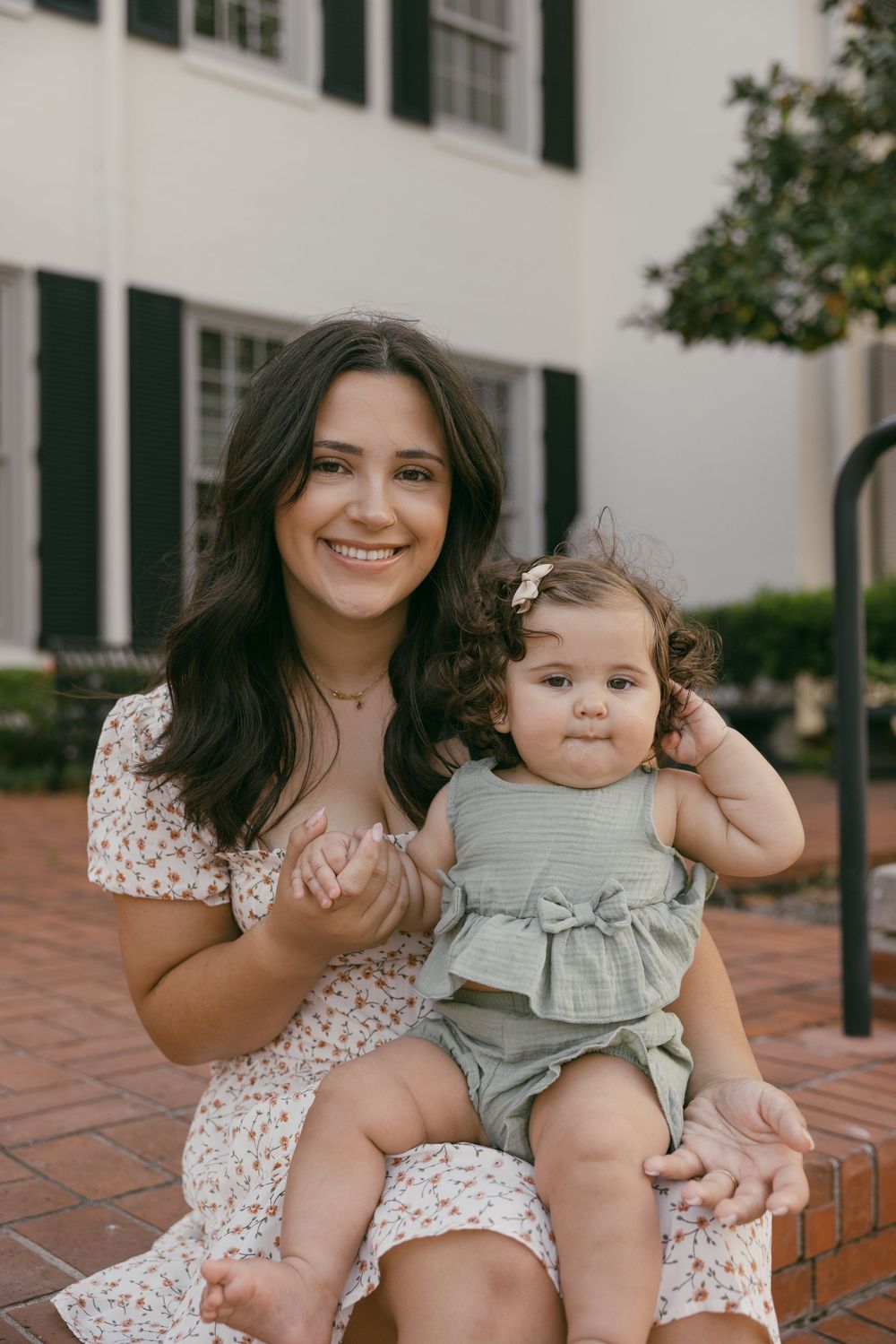 A woman in a floral dress sits with a baby wearing a gray romper on brick steps outside a white building.