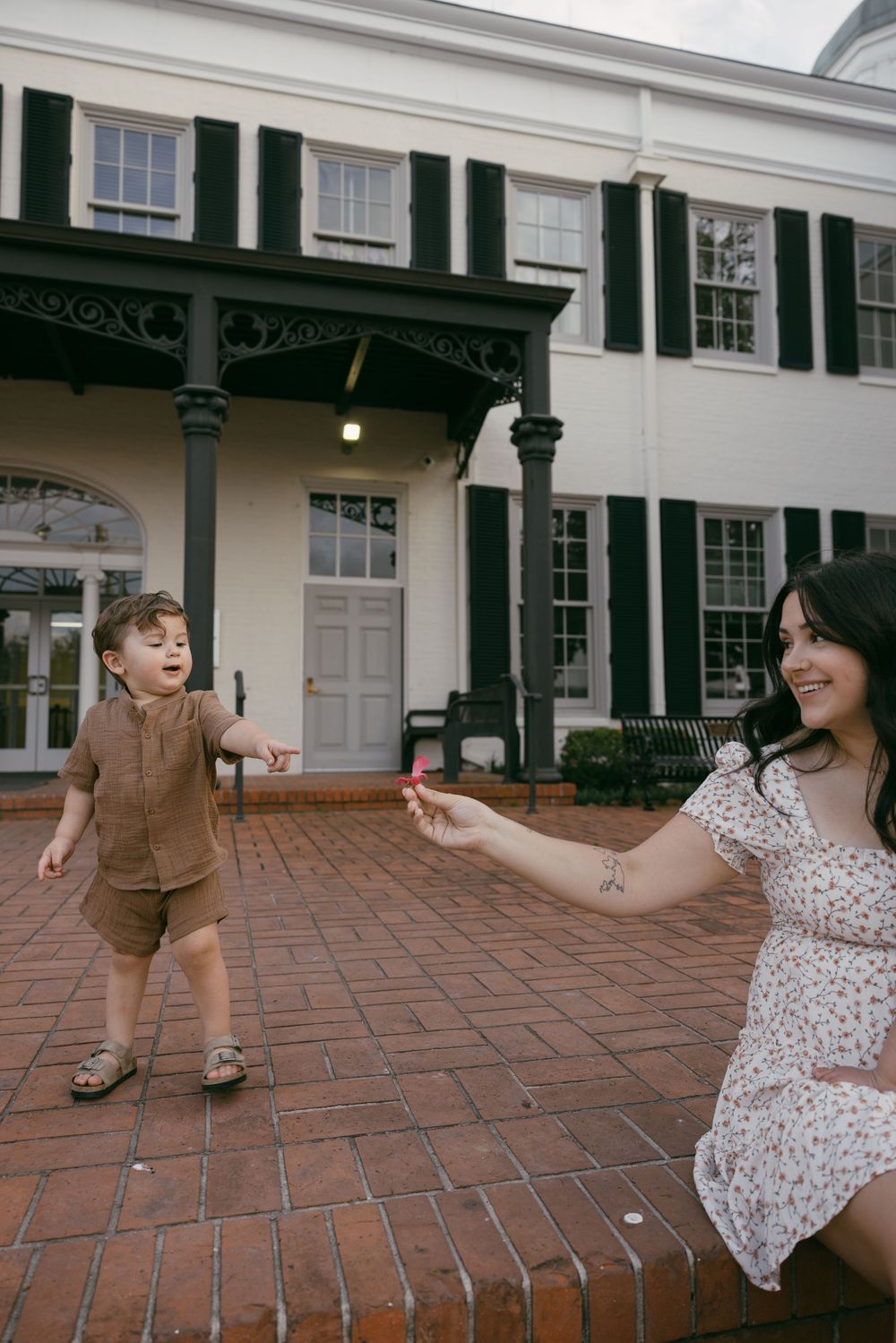 Person in a floral dress and boots walks along a brick pathway outside a white colonial-style building with black shutters.