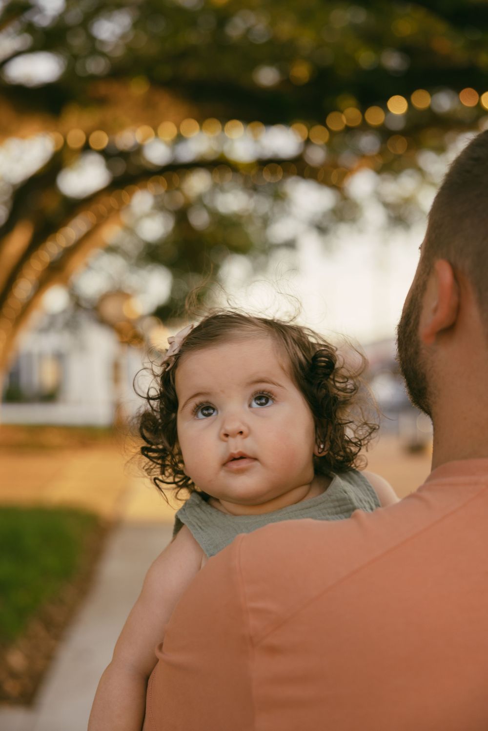 Baby with dark curly hair peers over a shoulder's shoulder in golden hour lighting at an outdoor location.