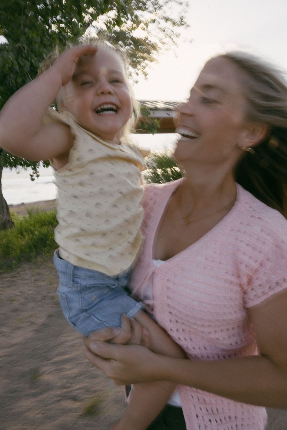 Woman in pink shirt playfully interacts with small child wearing patterned top against a sunny outdoor setting.