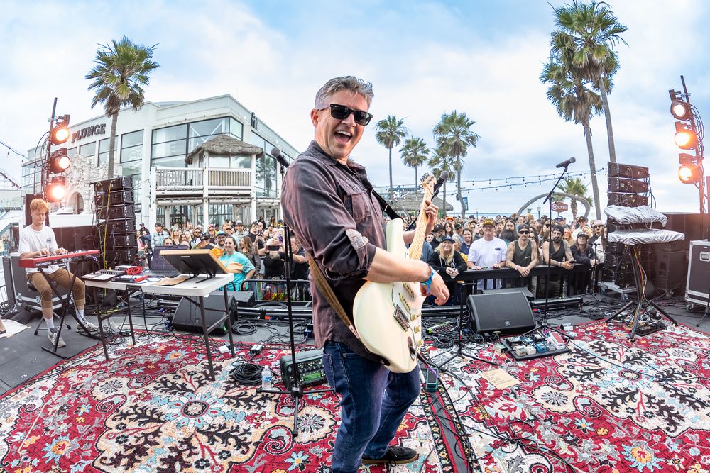 Guitarist smiling on an outdoor stage with palm trees and crowd at a sunny beachside music event.