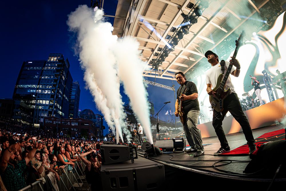 Two guitarists perform energetically on an outdoor concert stage with smoke effects and a large crowd at dusk.