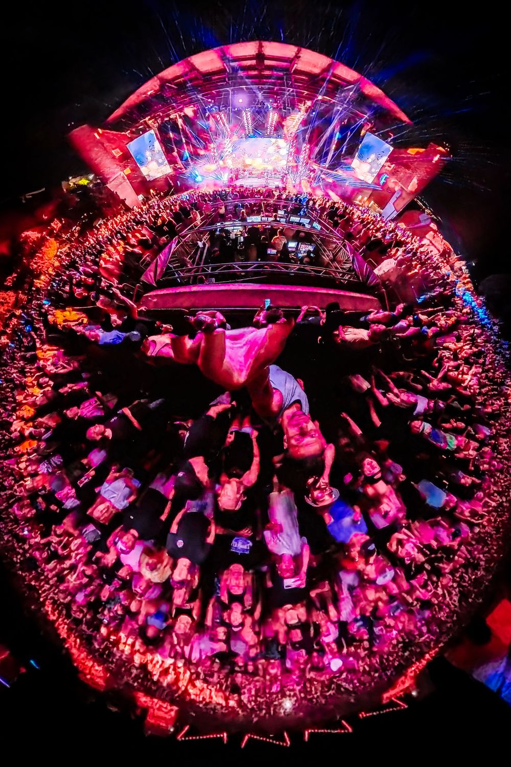 Fisheye lens view of a massive outdoor music festival crowd bathed in vibrant pink and red lights.