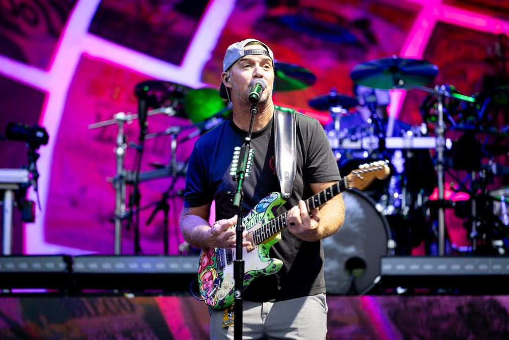 Male guitarist singing on stage with colorful guitar, wearing cap and black shirt, with drum kit visible behind him.
