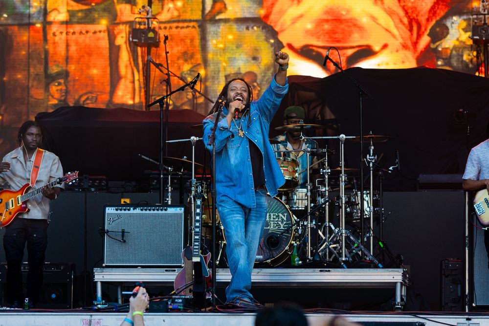 Energetic performer in blue jumpsuit singing on outdoor festival stage with band and colorful backdrop.