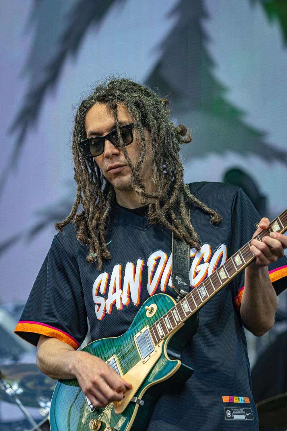 Guitarist with dreadlocks and sunglasses playing a teal Les Paul guitar on stage wearing a San Diego jersey.
