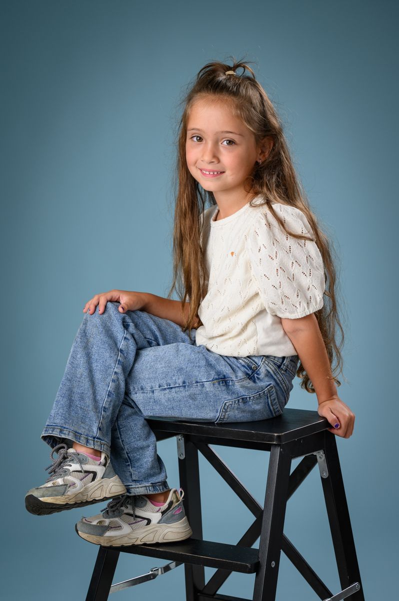 Child in white sweater and jeans sitting casually on black stool against blue background.