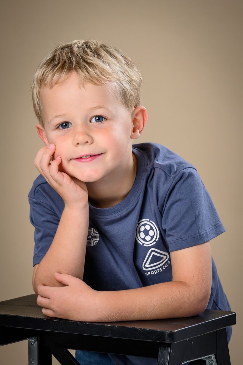 Child in blue t-shirt with patches resting head on hand against beige backdrop.
