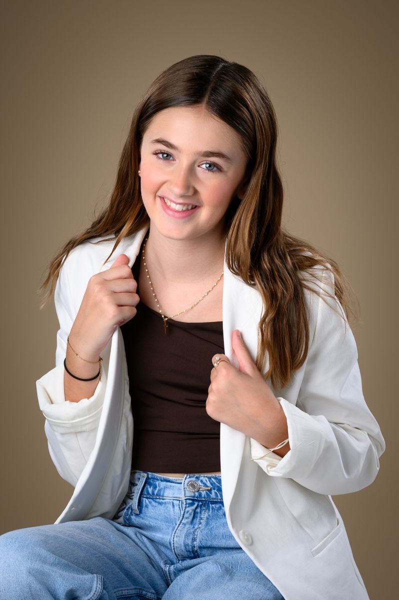 Young person in white blazer and brown top poses playfully against neutral backdrop.