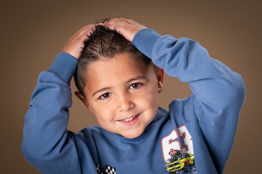 Child in blue sweatshirt makes playful poses against brown studio background.