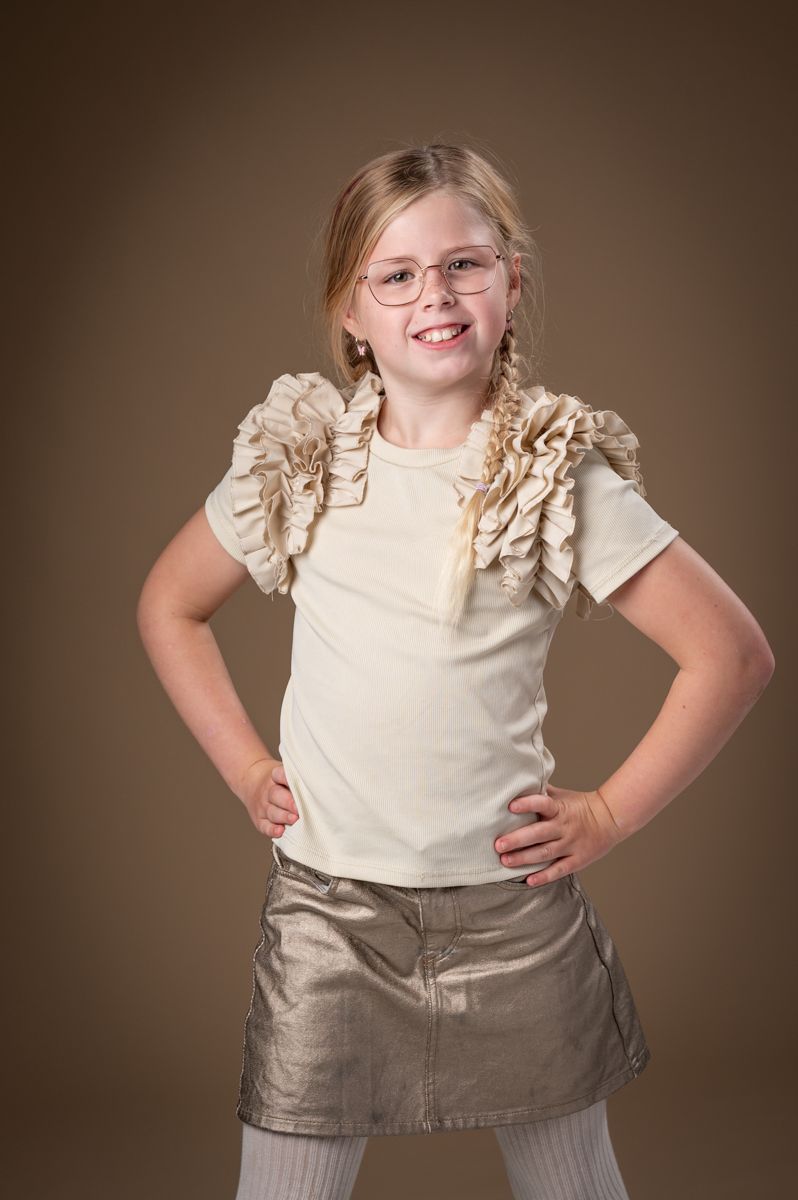Child wearing cream ruffled top and metallic skirt poses confidently against brown backdrop.