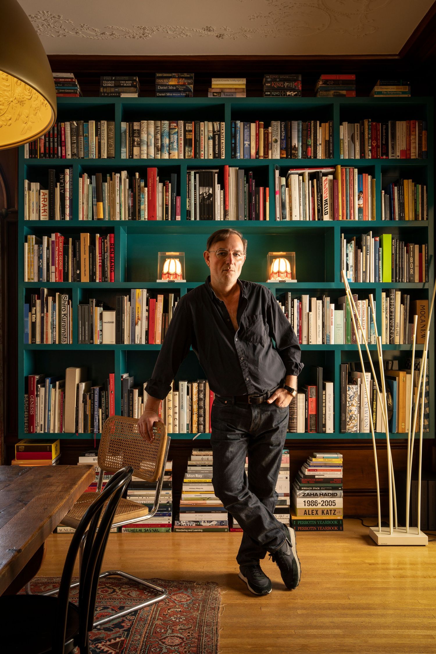 Man in black outfit leaning against teal bookshelf filled with books in a stylish home library.