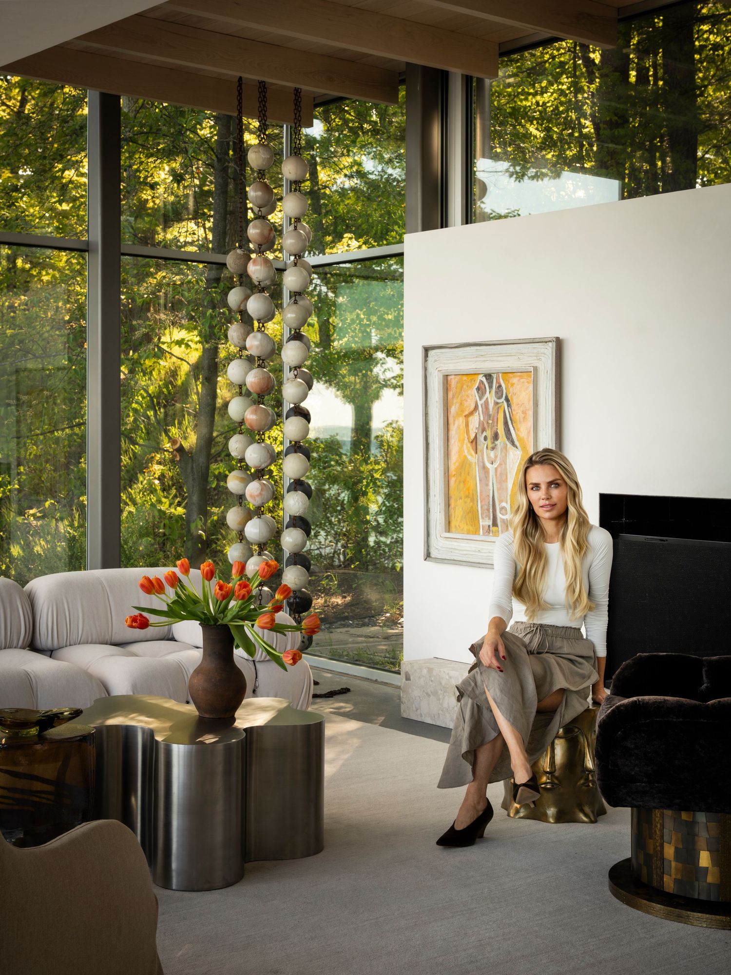 Woman sitting in modern living room with floor-to-ceiling windows, fireplace, abstract art, and sculptural pendant light.