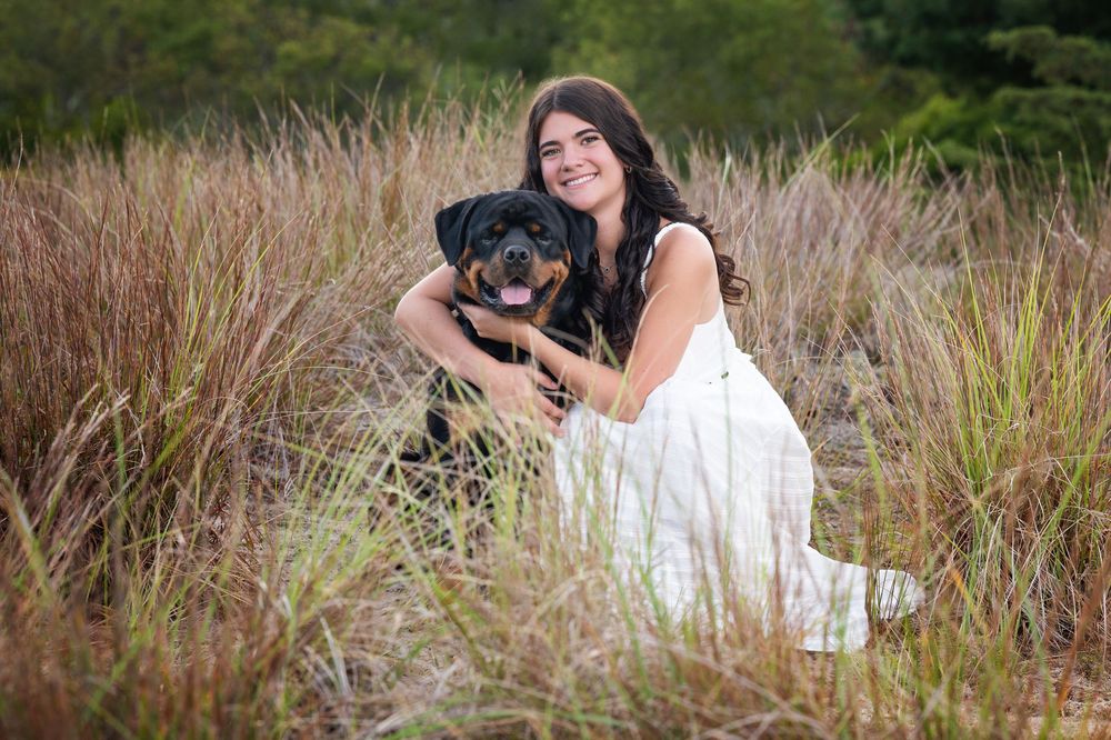 A person in white dress sits in tall grass with a black dog during an outdoor photoshoot.