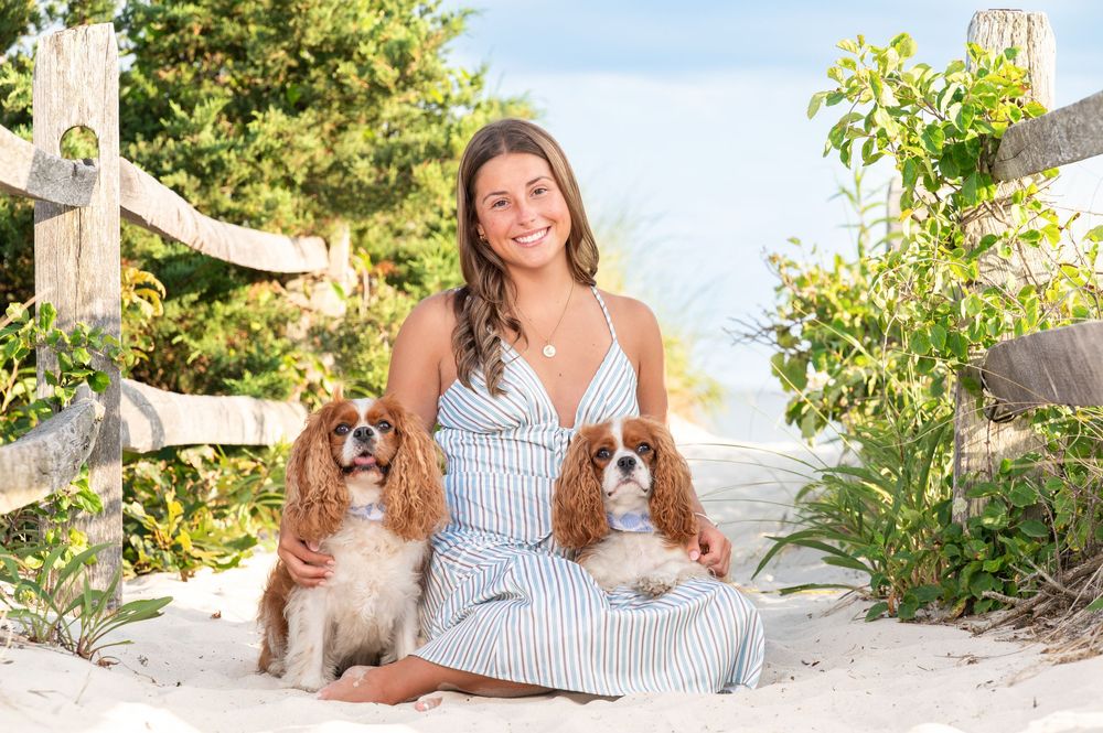 Two Cavalier King Charles Spaniels sit outdoors with someone in a striped dress on a sunny summer day.