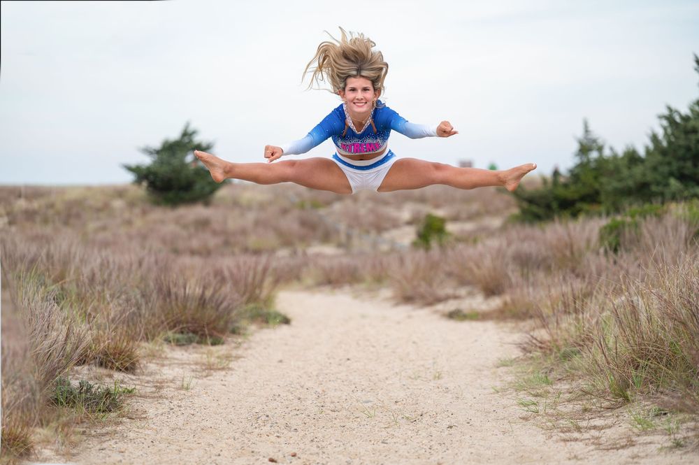 A person joyfully leaps across a sandy beach path through coastal vegetation on a sunny day.