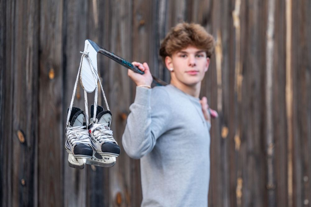 Person in gray sweater holds up ice skates in front of rustic wooden wall.