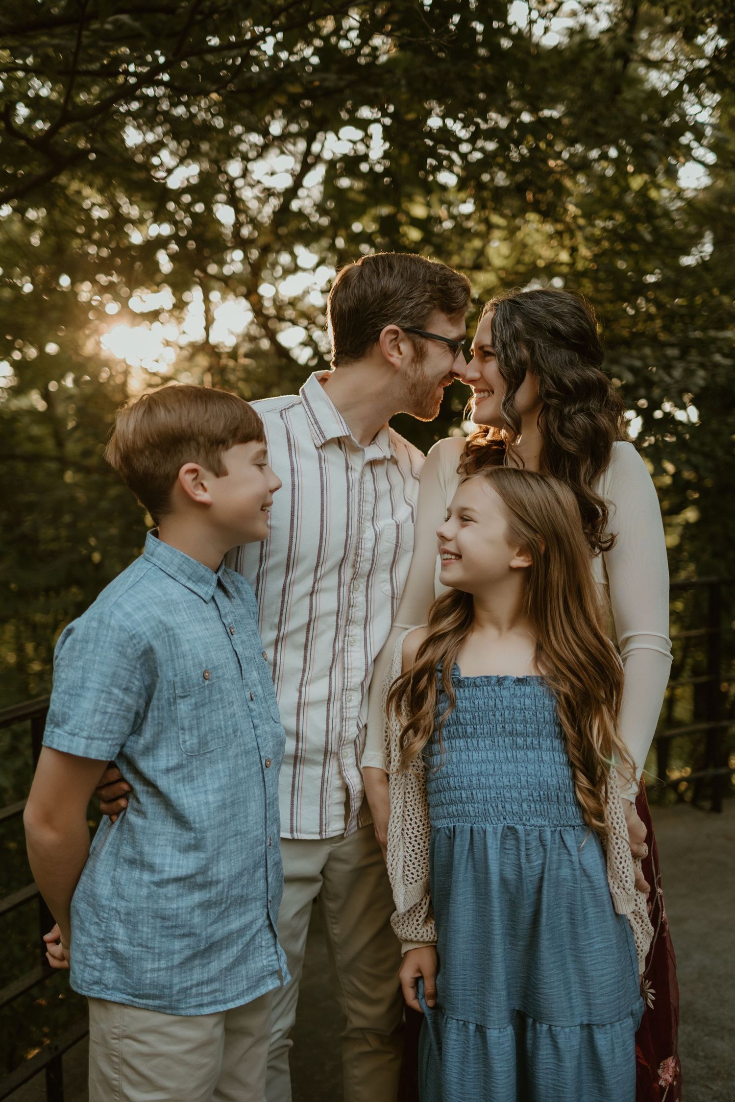 A family poses together outdoors during golden hour with sunlight filtering through trees in the background.