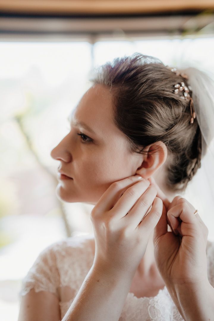 A bride wearing a delicate lace dress and pearl hair accessories puts in her earring during wedding preparations.