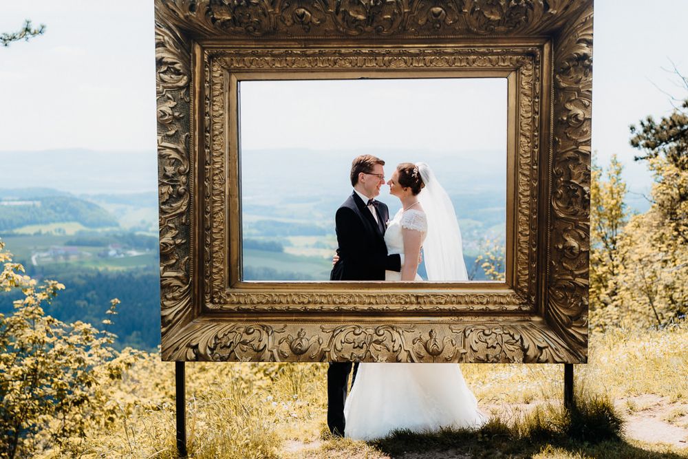 Newlyweds stand behind an antique gold frame on a hilltop overlooking a valley.