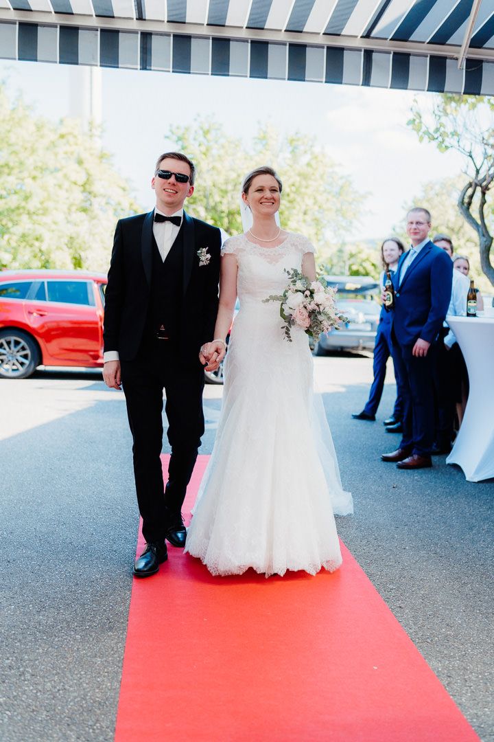 Wedding couple walking down red carpet under striped awning with guests watching.