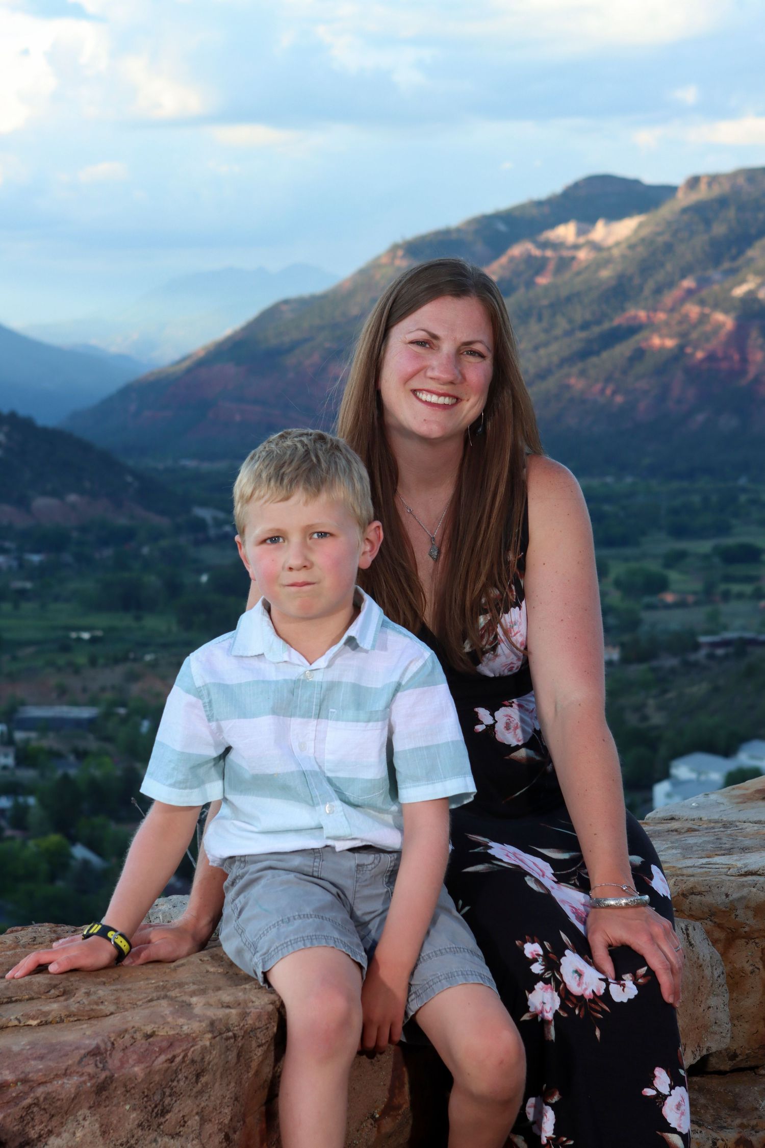 A pair sits together on a rock overlook with scenic red rock mountains and valley views behind them at sunset.