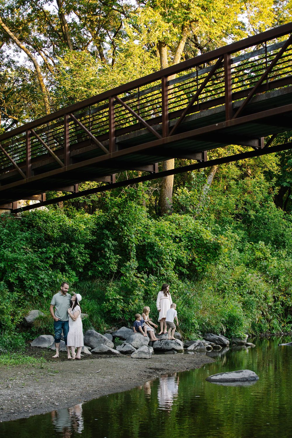 A peaceful scene under a rustic bridge with people sitting by the water's edge.
