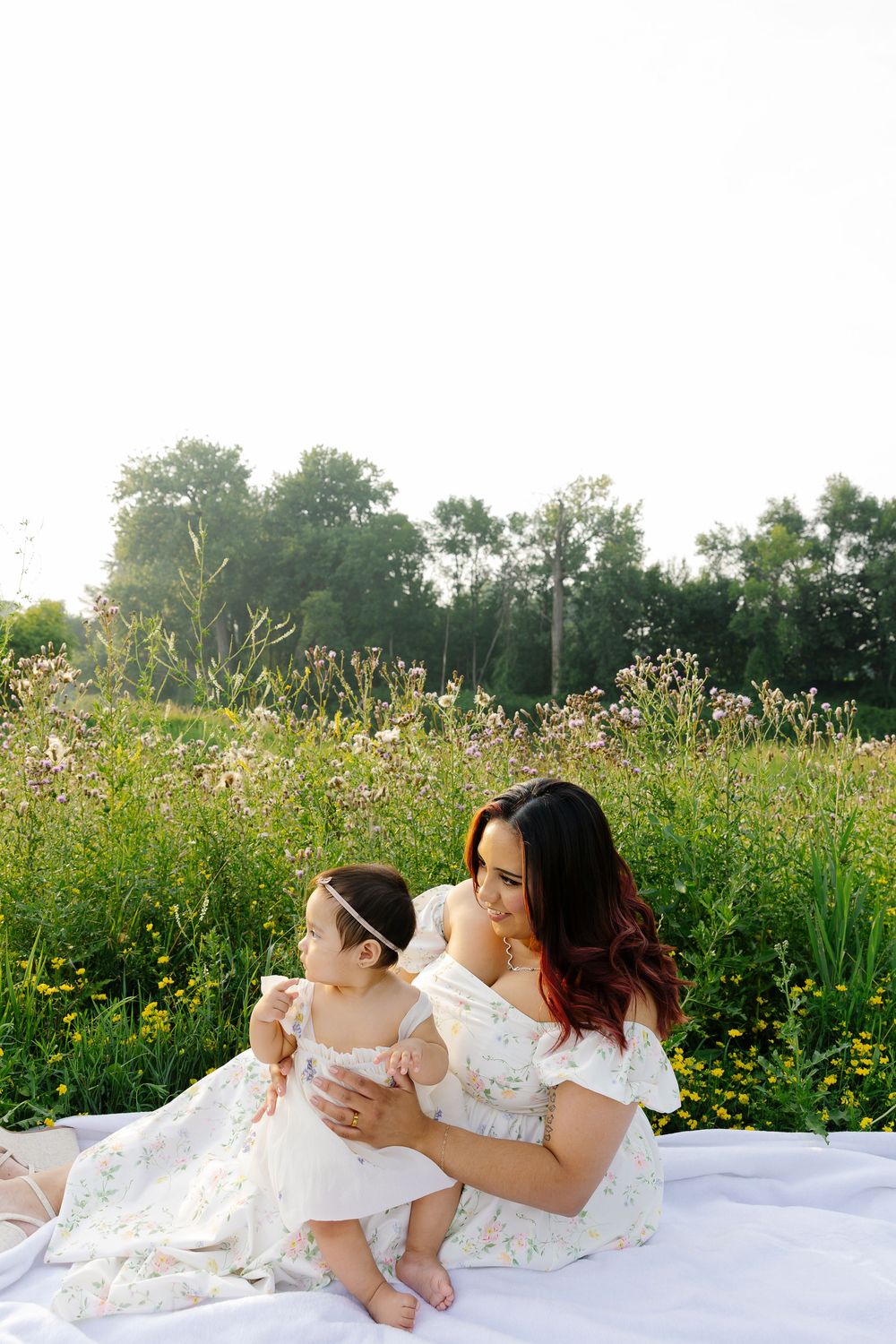 Mother and baby in white dresses sit on blanket in wildflower meadow during golden hour.