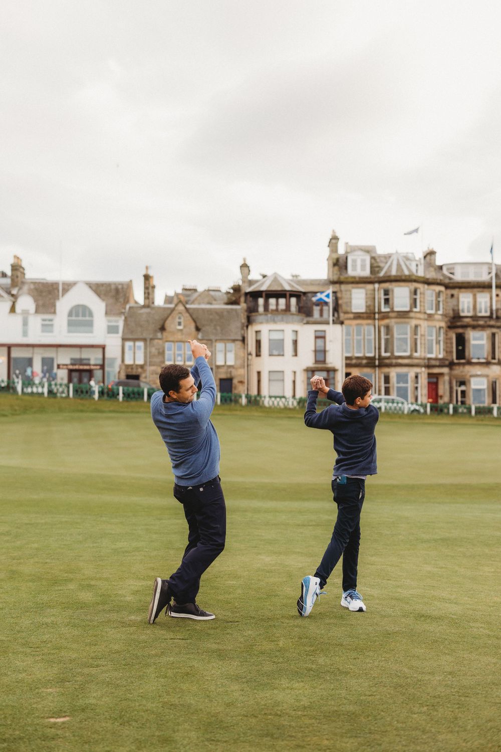 Two golfers practicing their swings on a course with traditional Scottish buildings in the background.