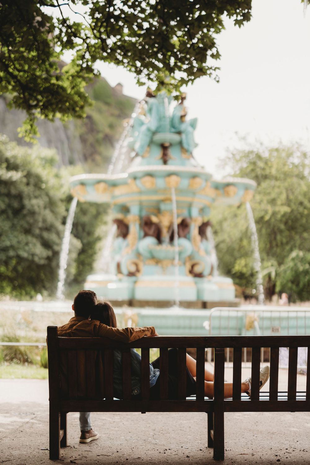 A couple sits on a bench overlooking an ornate turquoise fountain in a park setting.
