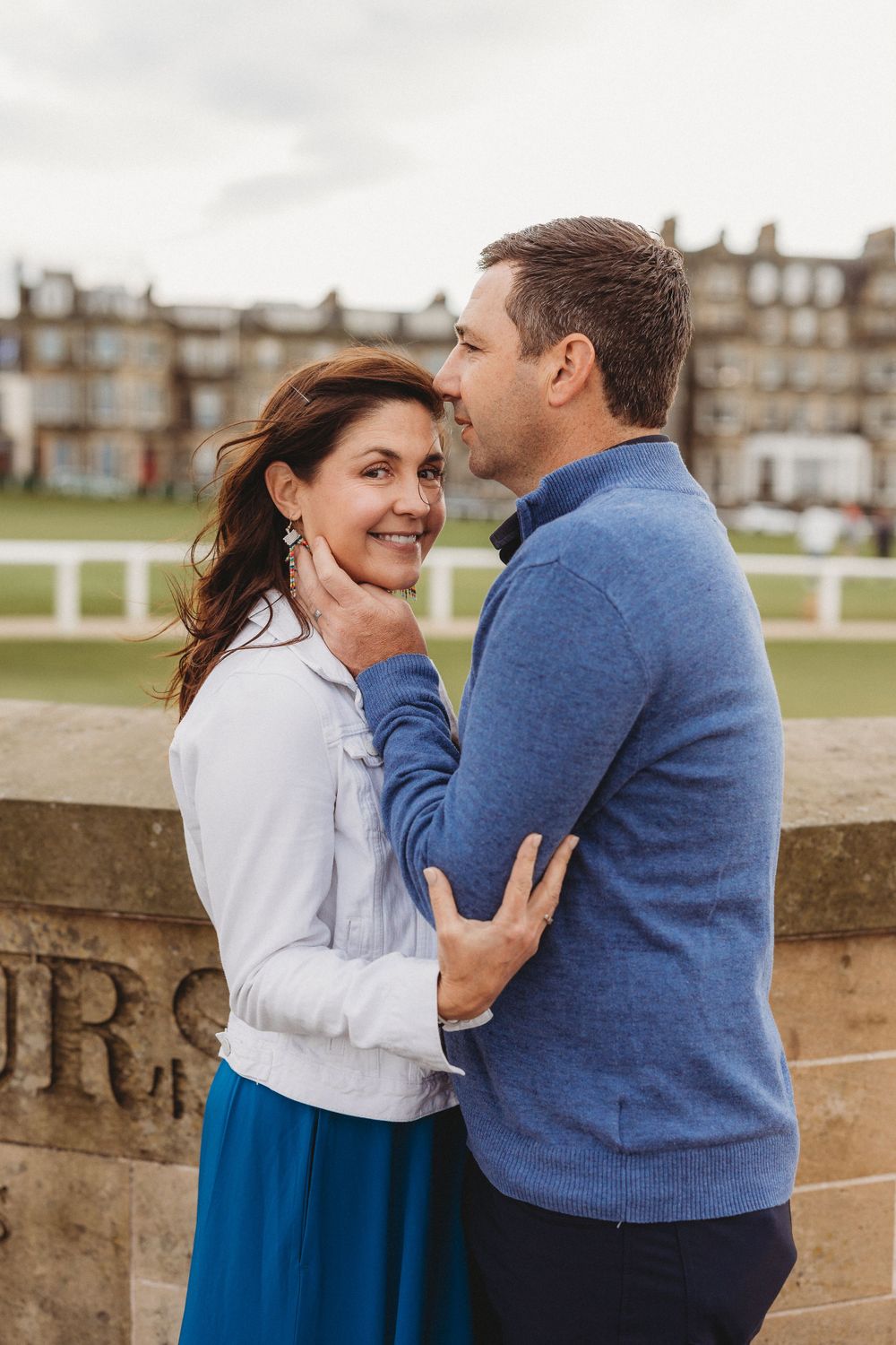 A couple embraces on a stone wall with historic buildings in the background.