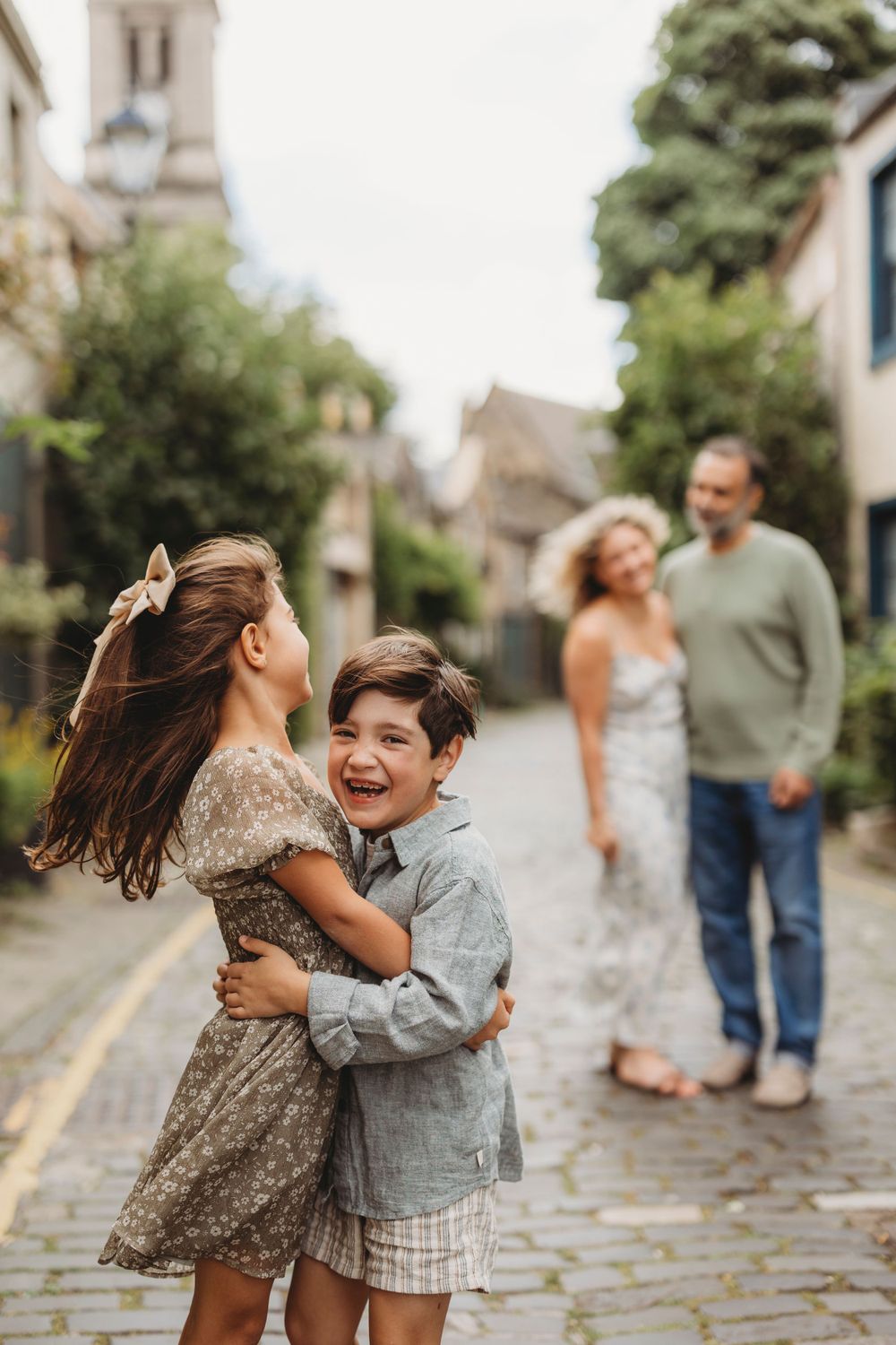 Two children hugging and laughing on a cobblestone street with adults in the background.