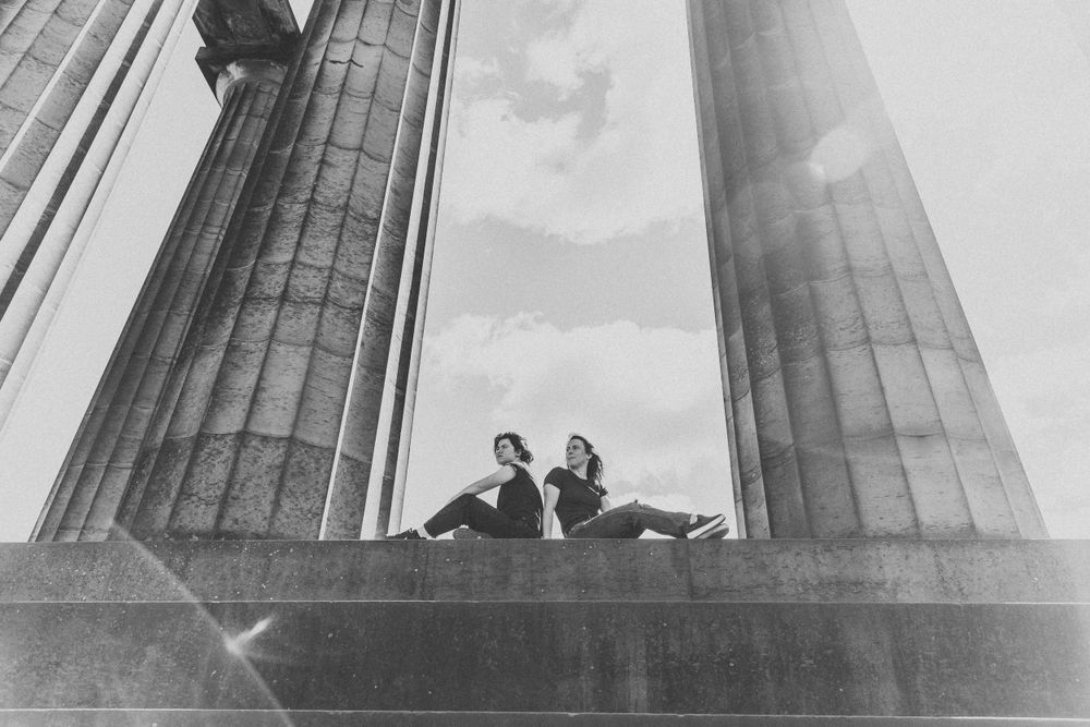 Two figures sit between massive classical columns against a misty sky in black and white.