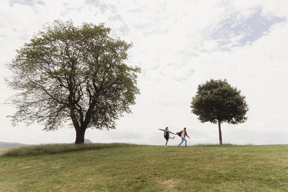 Two silhouettes dance playfully between two trees on a grassy hilltop against a cloudy sky.