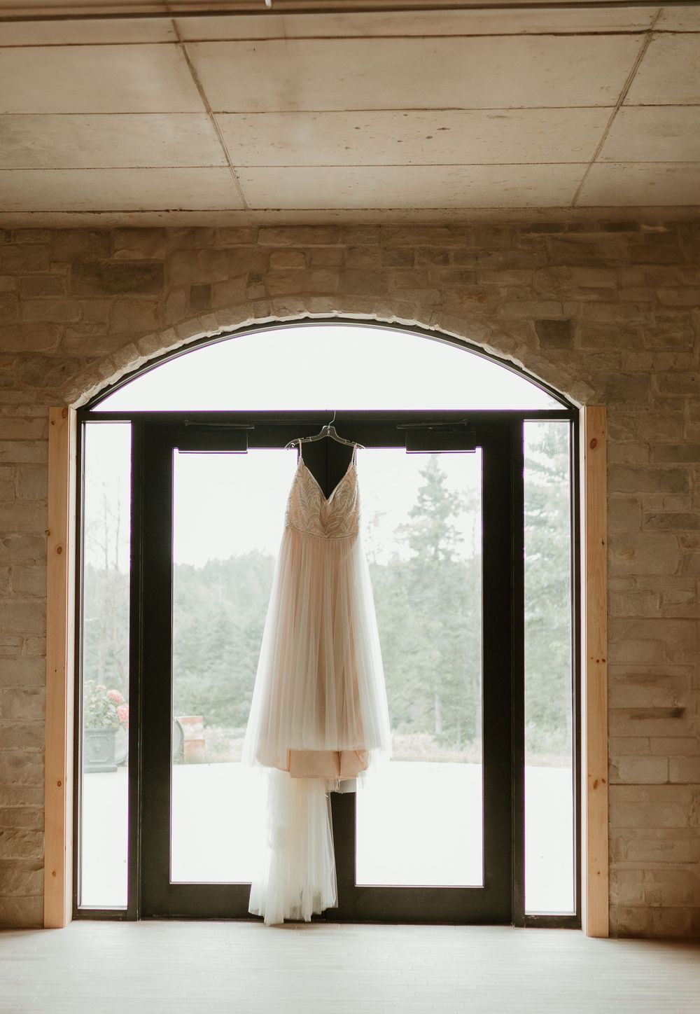 Elegant white wedding dress hanging in arched doorway with natural light streaming through glass doors.