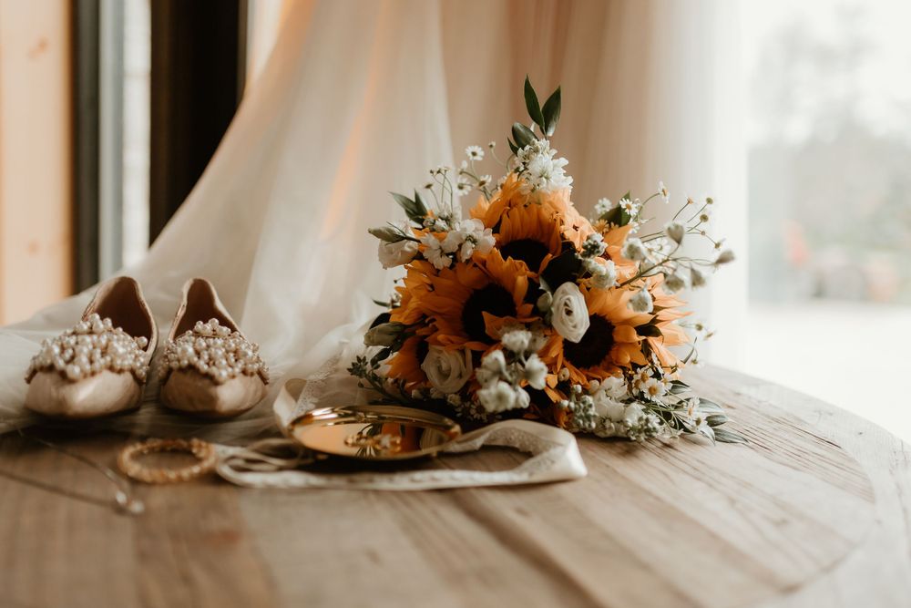 Bridal flat lay featuring sunflower bouquet, pearl shoes, and gold jewelry on a wooden table.