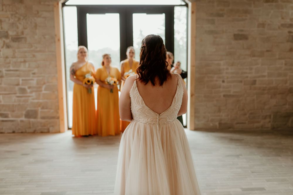 Bride in lace wedding gown faces bridesmaids in yellow dresses for emotional first look reveal.