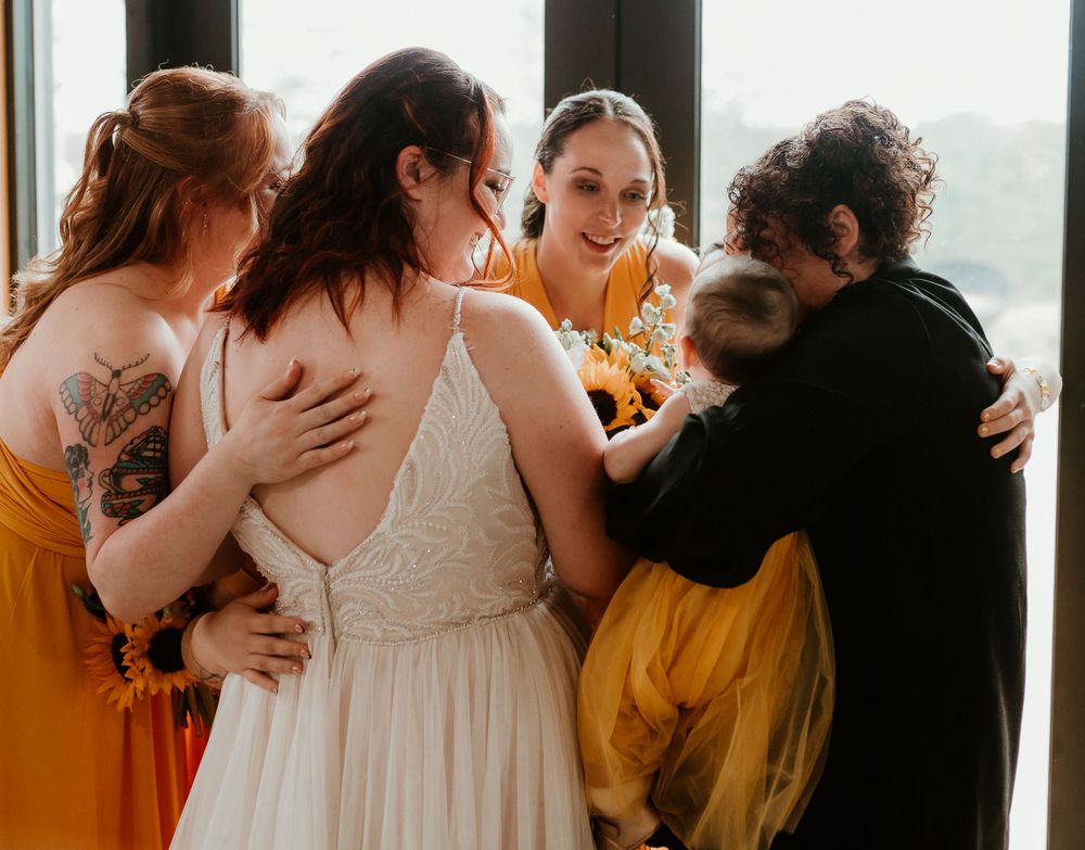 Bride surrounded by bridesmaids in orange dresses sharing a joyful group hug near a window.