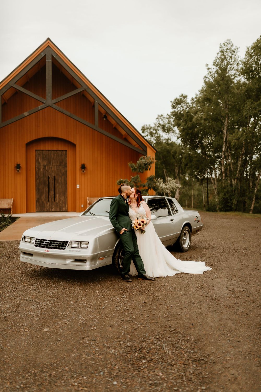 Bride and groom kissing beside a vintage silver car in front of a rustic wooden chapel.
