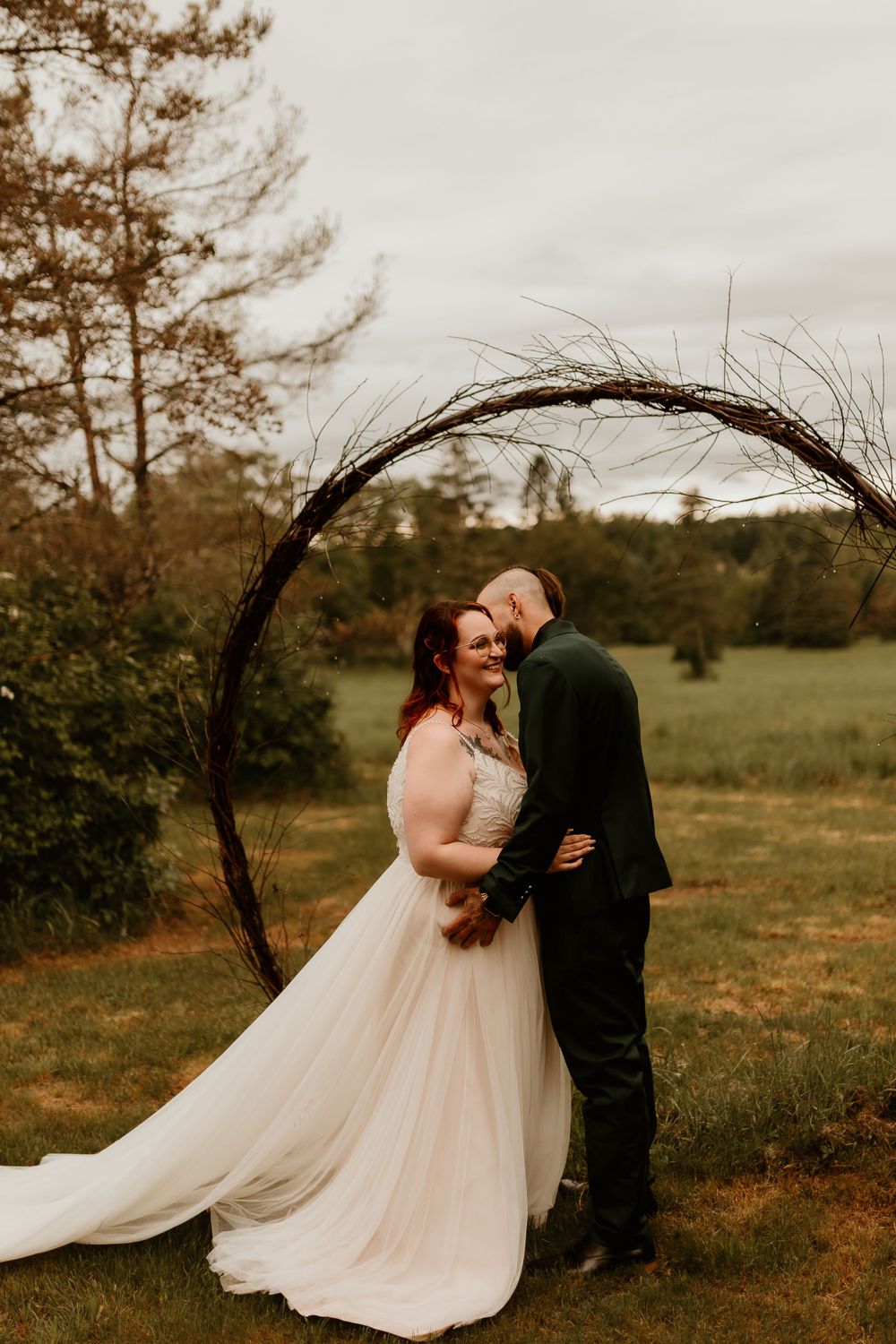 Bride and groom embracing under a circular twig arch in a lush green outdoor wedding setting.